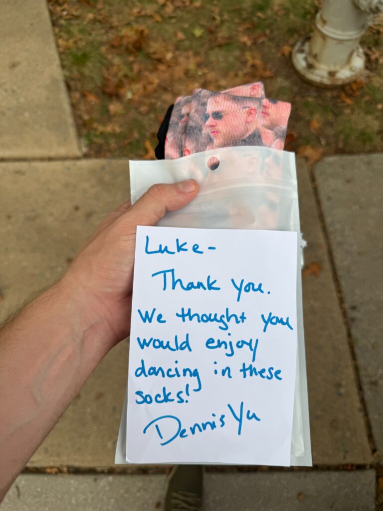 Luke holding personalized socks with his face on them and a handwritten note from Dennis Yu thanking him, set against a sidewalk background.