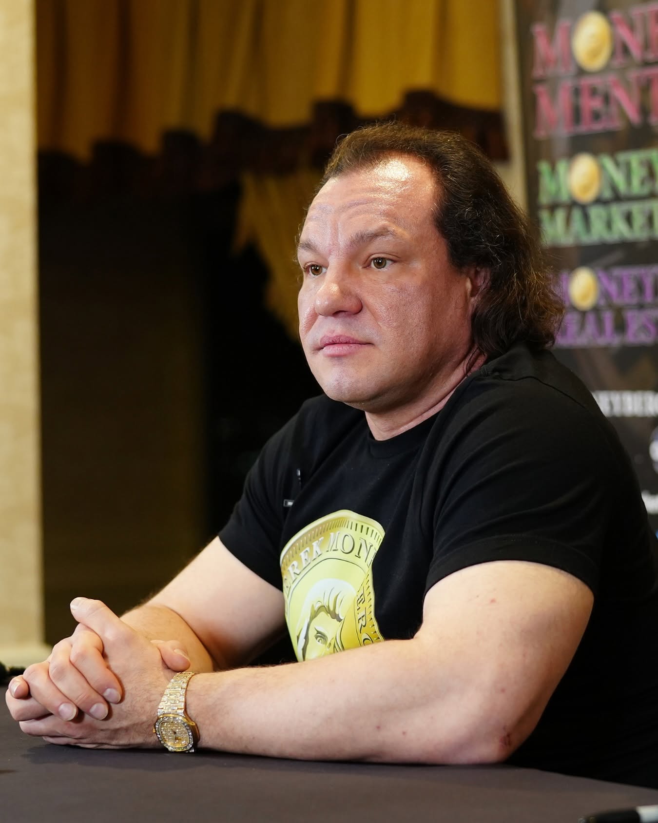 Derek Moneyberg seated at a table wearing a black and gold Moneyberg shirt.